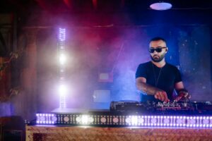 A DJ wearing sunglasses mixing music on a deck with vibrant lights and smoke in an indoor club.