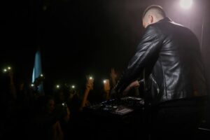 DJ in black leather jacket energizes young crowd at night with vibrant music and lights.